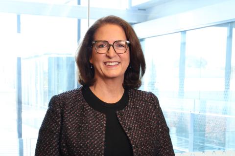 ACIC CEO Heather Cook smiles at the camera. She has short brown hair and wears glasses. She is stnading in front of glass wall. 
